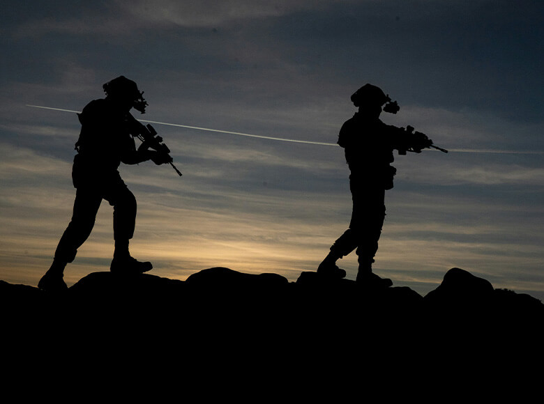 Silhouettes of two people in tactical gear walking across rocky terrain at dusk, each holding a long firearm and wearing head‑mounted night‑vision equipment.