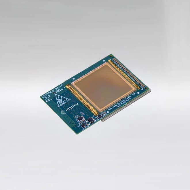A square microdisplay module mounted on a green printed circuit board. The center features a large reflective display area bordered by fine gold contact traces. Surrounding the display are various electronic components, connectors, and printed markings, including the Kopin logo and circuit identifiers. The board is shown on a light gradient background.
