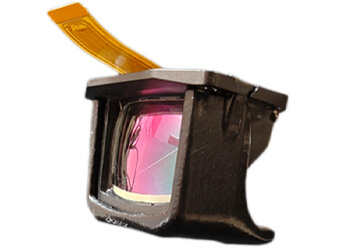 A compact, rectangular augmented reality optical module with a black housing and a reflective, pink‑tinted see‑through lens on the front. A thin, curved yellow flex cable extends from the top of the unit. The device is shown isolated on a white background and represents Kopin’s DayVAS clip‑on AR display system.